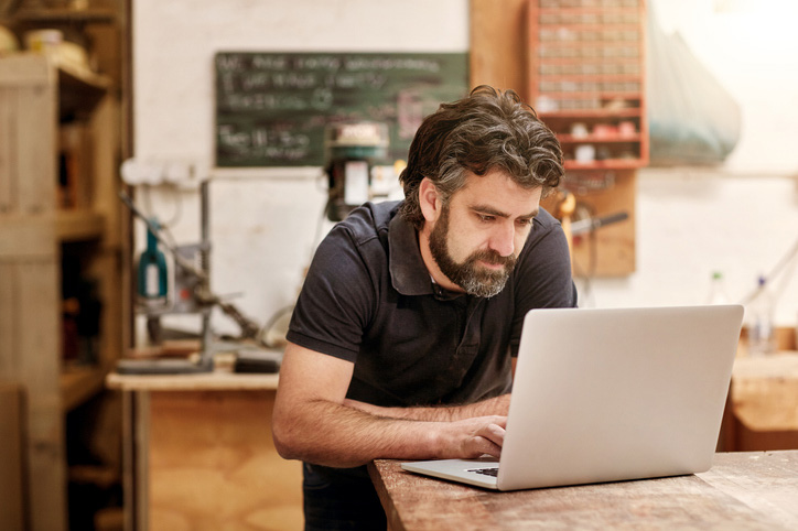 a person stands, resting on a workbench, looking at a laptop
